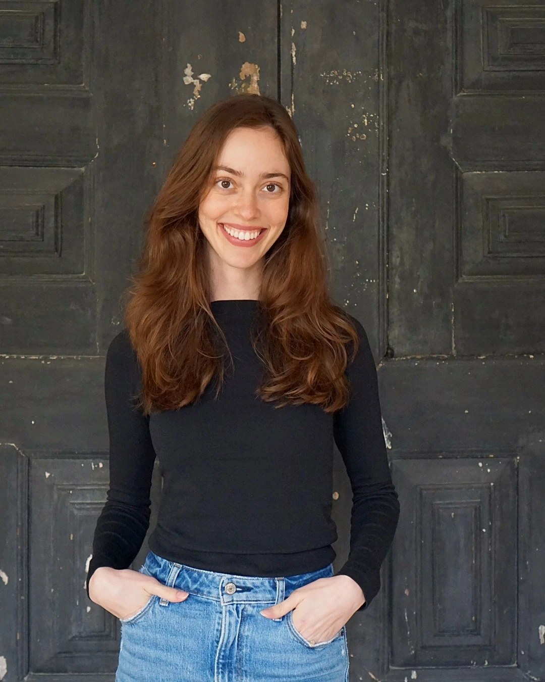 Portrait of Leonor, founder of Enleio Events, smiling in a black top and jeans against a weathered dark wooden door