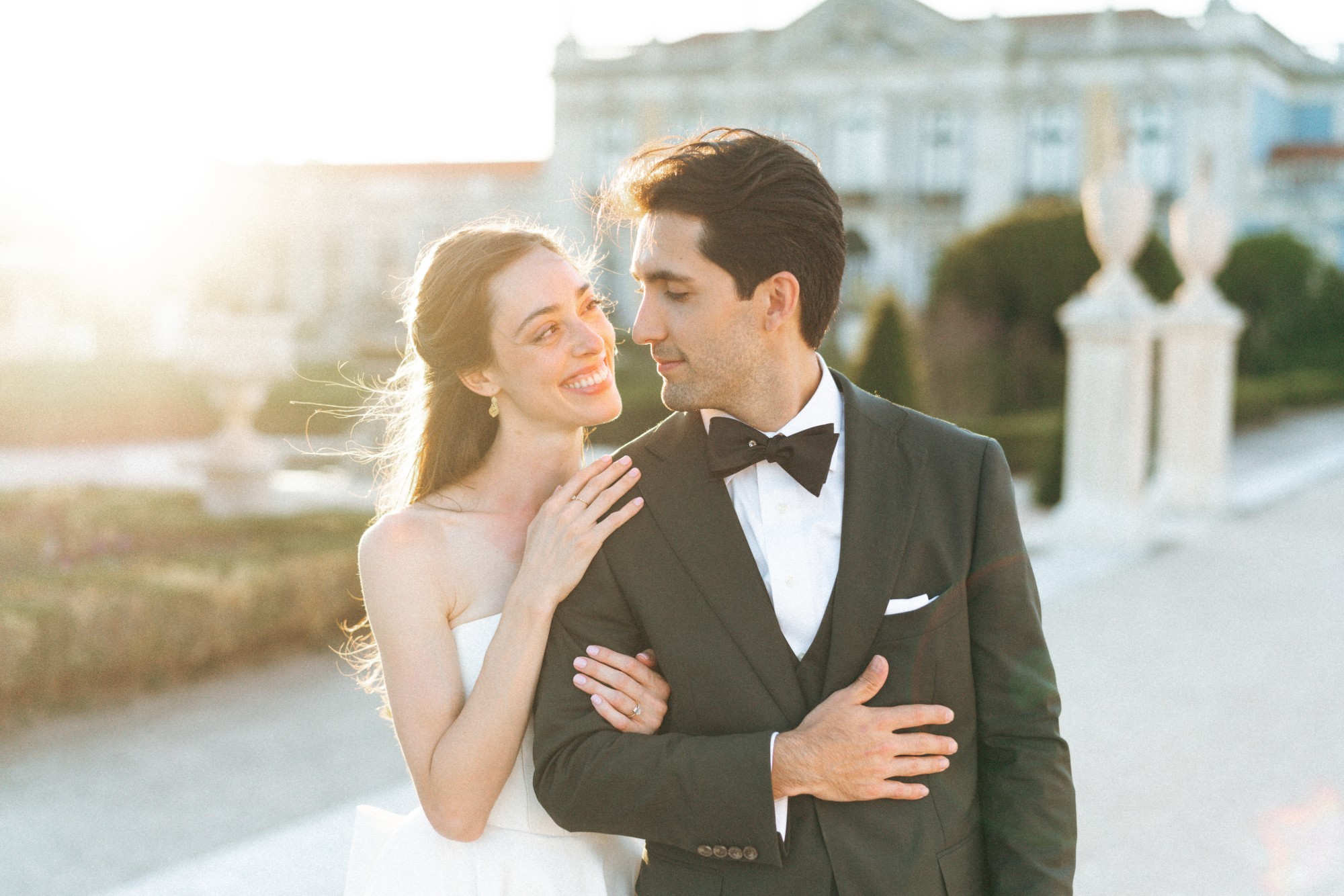 A smiling bride rests her hand on her groom's shoulder as they look at each other in the golden light, with the Palácio Nacional de Queluz behind them