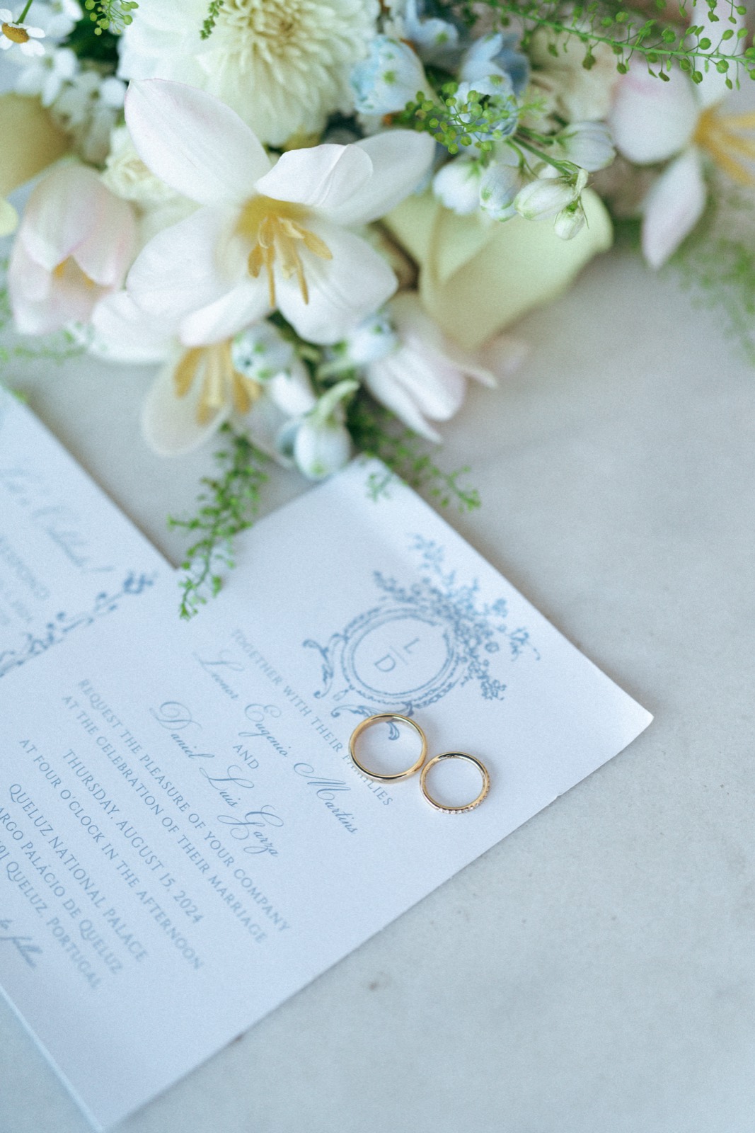 Wedding invitation with a blue crest, with two gold rings resting on it and white flowers above