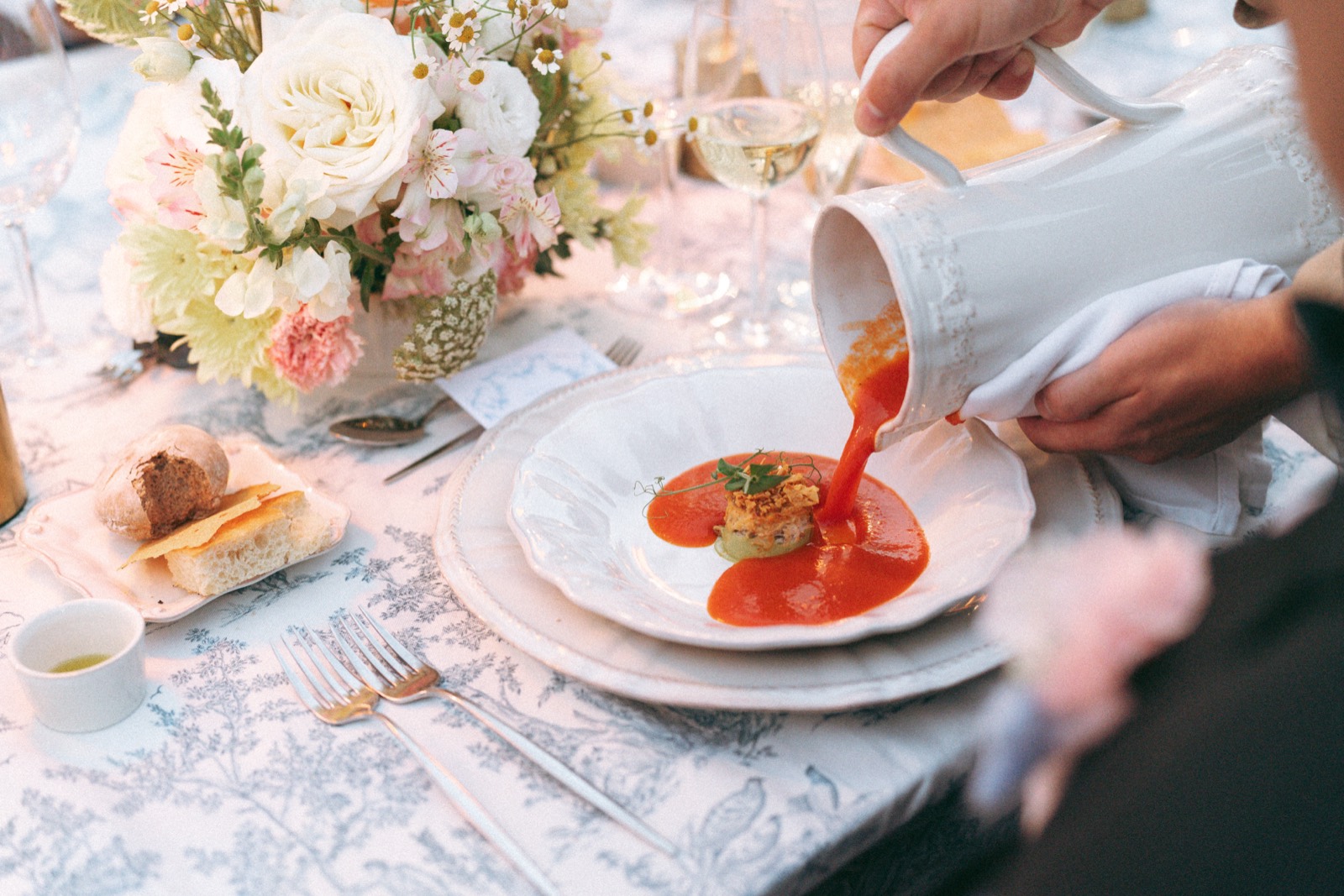 A server pouring red soup from a white pitcher into a plate on a blue toile tablecloth