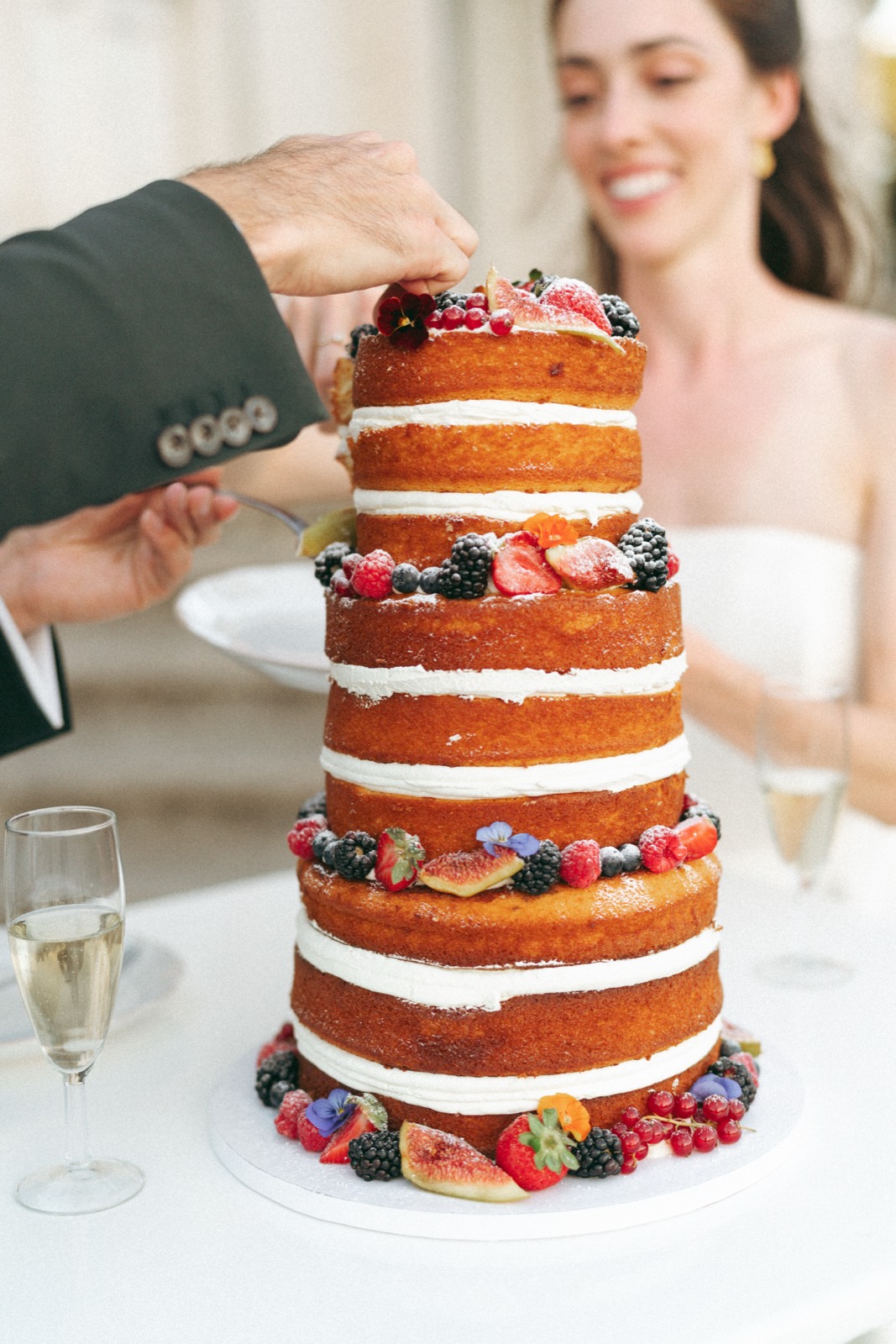 A tall naked cake with berries and figs being cut, with the bride smiling behind