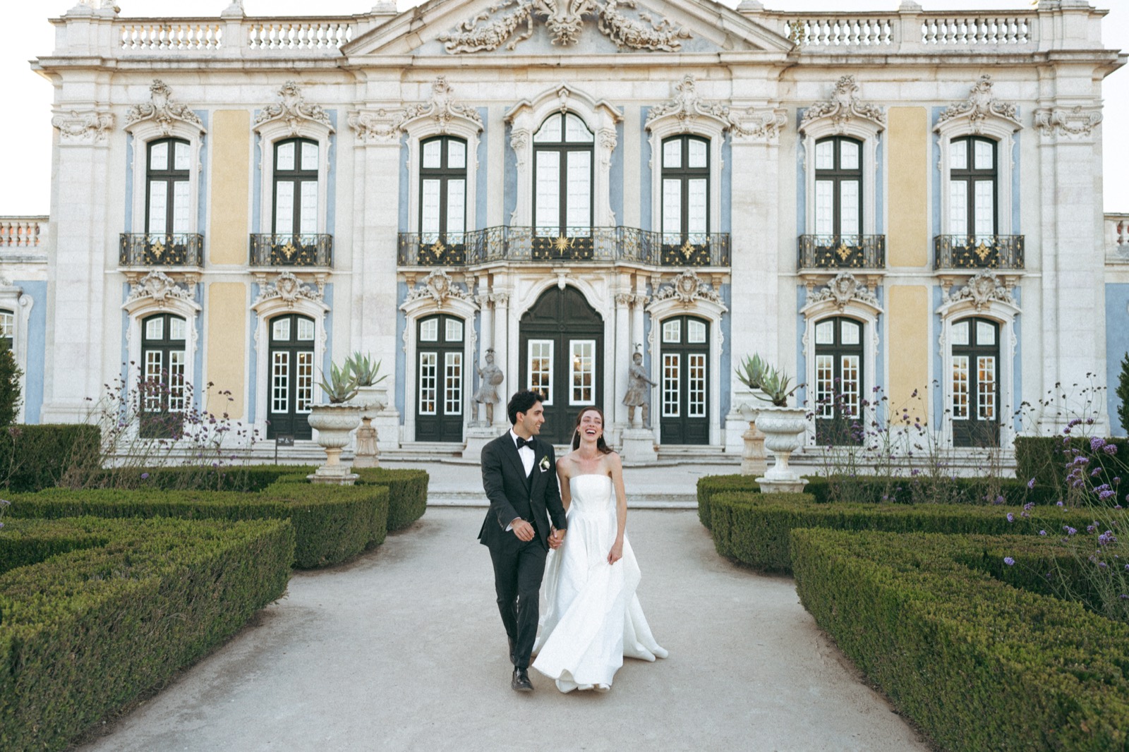 The couple laughing as they walk toward the camera in front of the palace