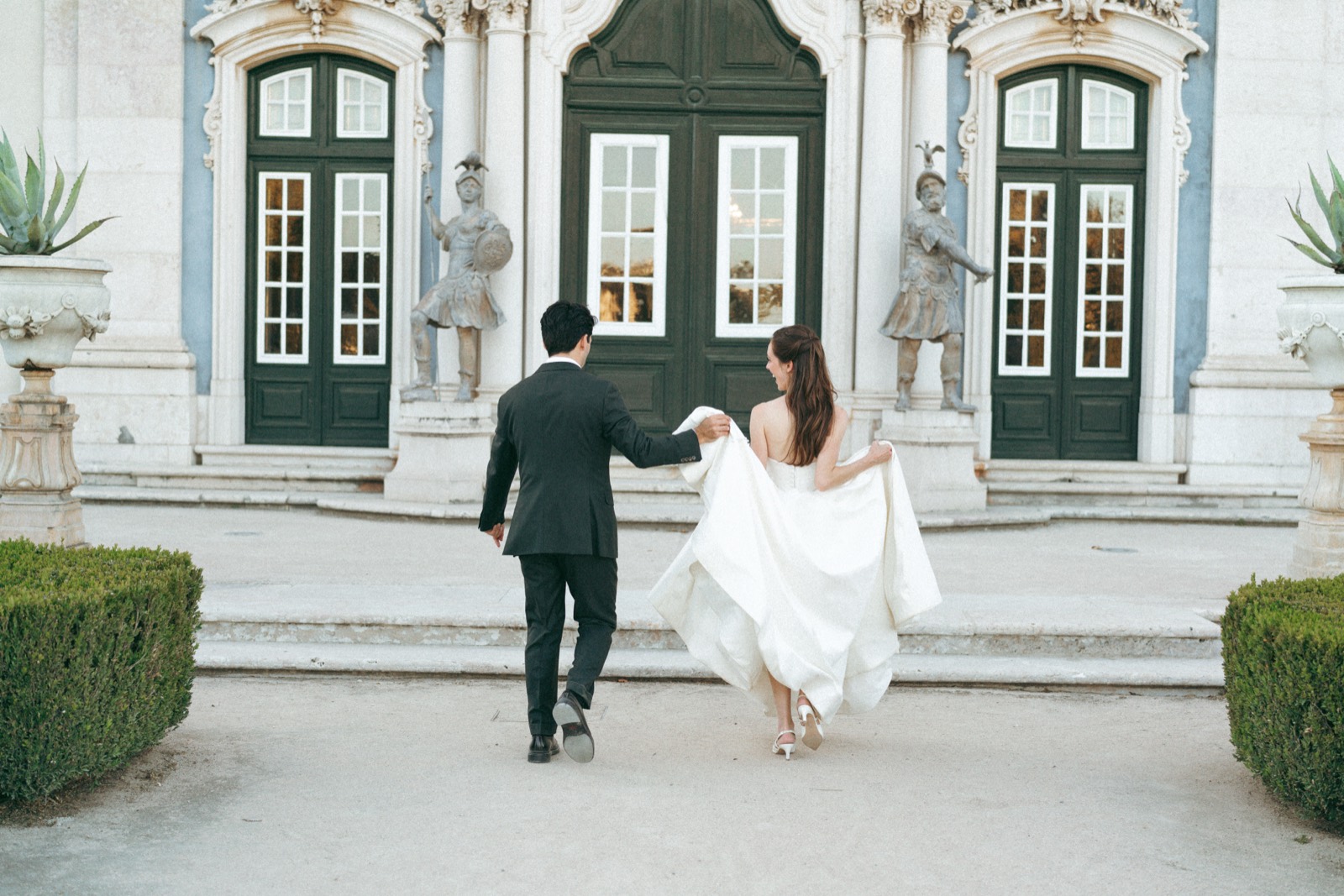 The couple walking away from the camera past the palace doors, the bride holding her gown