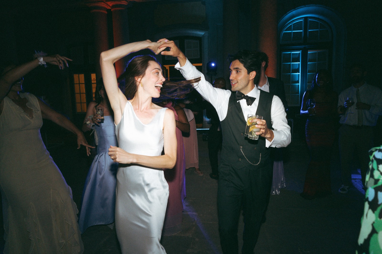 The bride laughing mid-twirl in a white slip dress as the groom spins her, with guests dancing around them