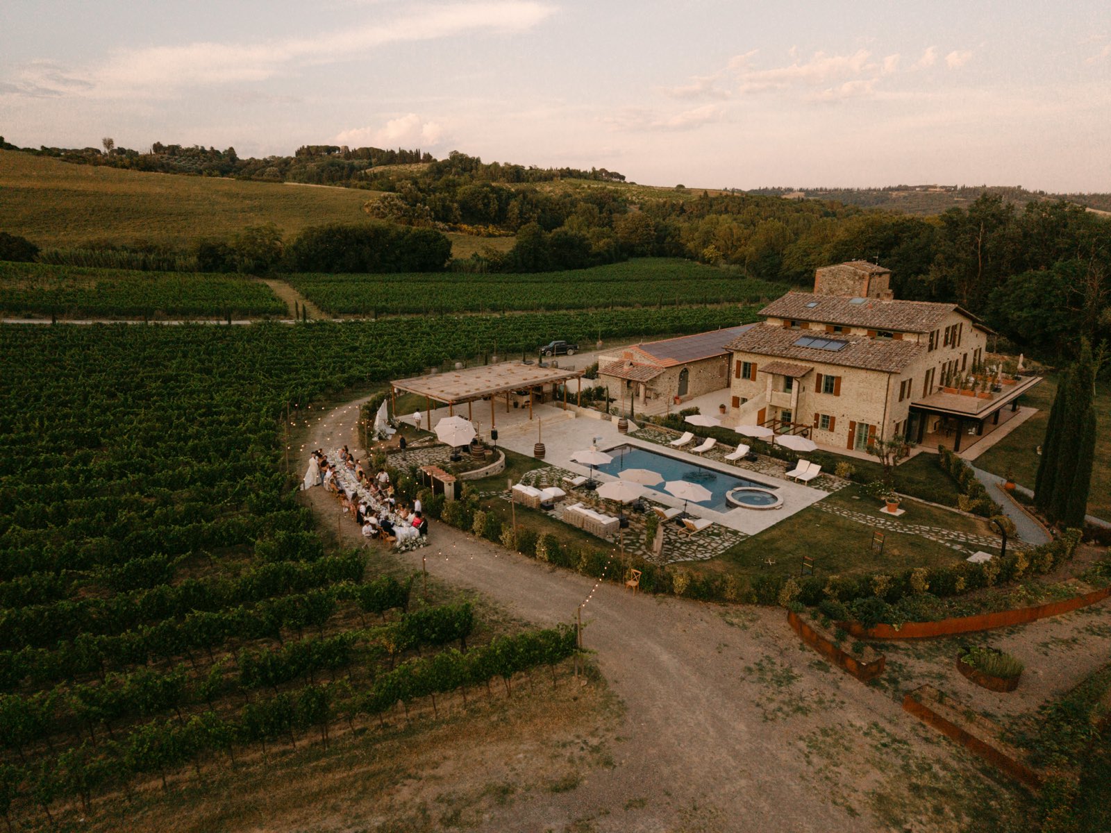Aerial view of a stone villa with a pool, and a long dinner table set along a curving path through the vineyard at dusk