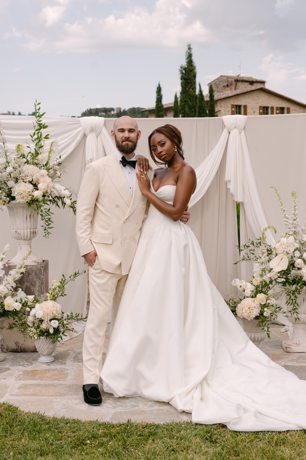 A smiling groom in a cream double-breasted suit stands beside his bride in a strapless satin gown, in front of draped ivory fabric and white florals