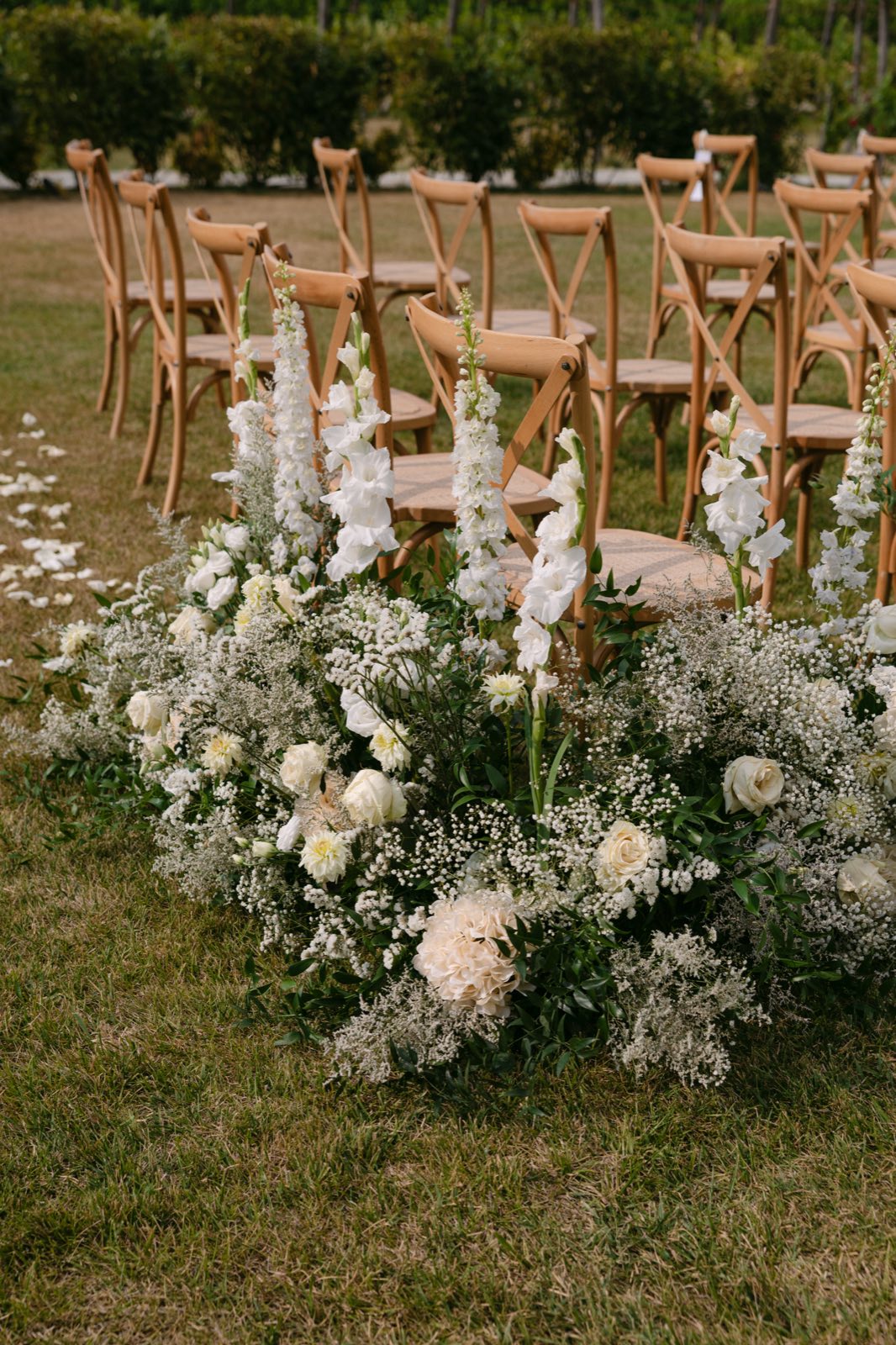 A ceremony aisle lined with chairs and a low arrangement of white delphinium, gladioli, roses and baby's breath on the grass
