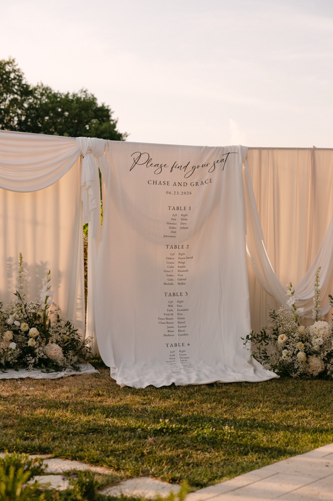 A white fabric seating chart hung between draped ivory curtains and flanked by white floral arrangements, reading Please find your seat