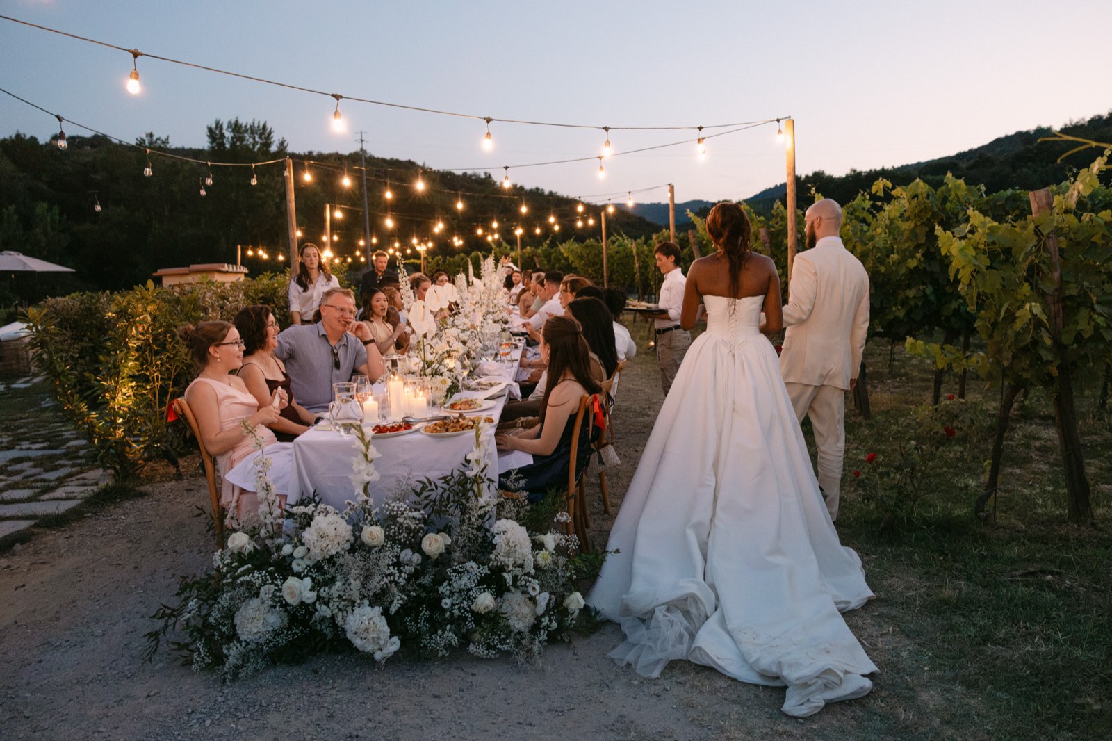Guests seated along a long vineyard table under string lights at dusk, as the couple walk past a low floral arrangement