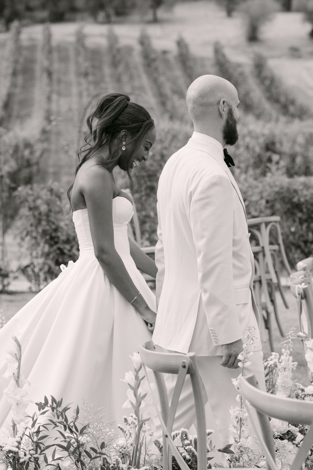 Black and white photo of the bride laughing as she walks hand in hand with her groom past rows of chairs and flowers