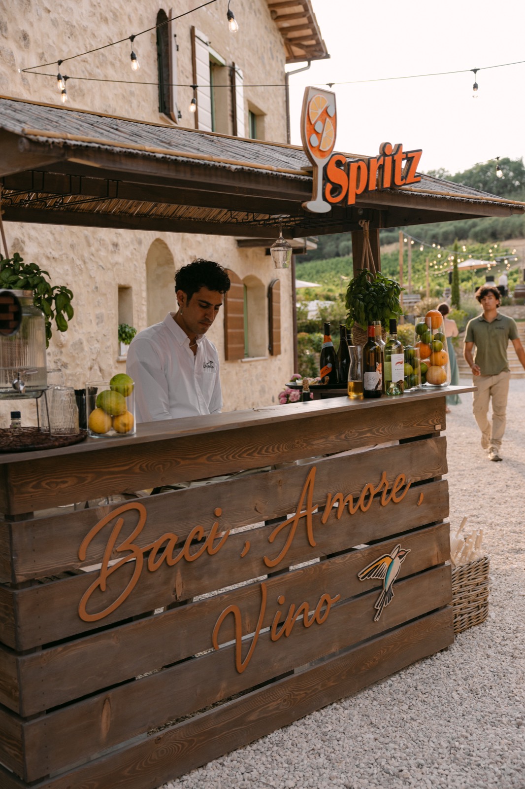 A wooden bar with a Spritz sign and carved lettering reading Baci, Amore, Vino, with citrus and basil on top and a bartender behind it