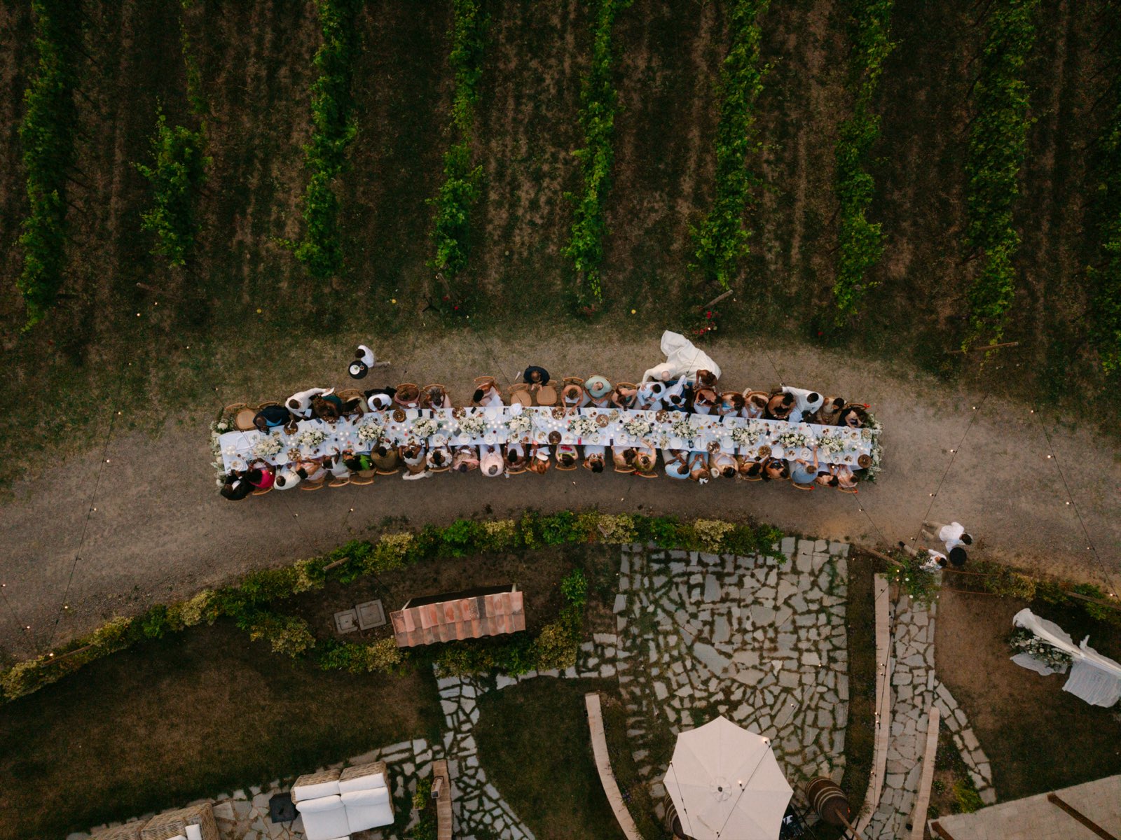 Overhead drone view of a long dinner table set with white linens and florals, running along a dirt path between vineyard rows