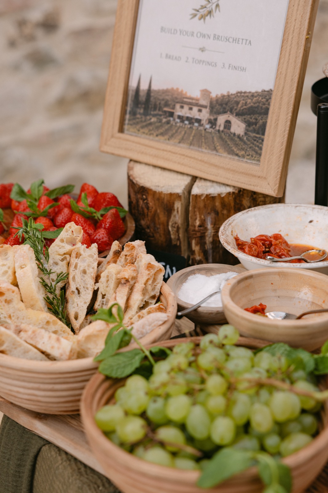 A build-your-own bruschetta station with sliced bread, strawberries, grapes and tomatoes beneath a framed sign