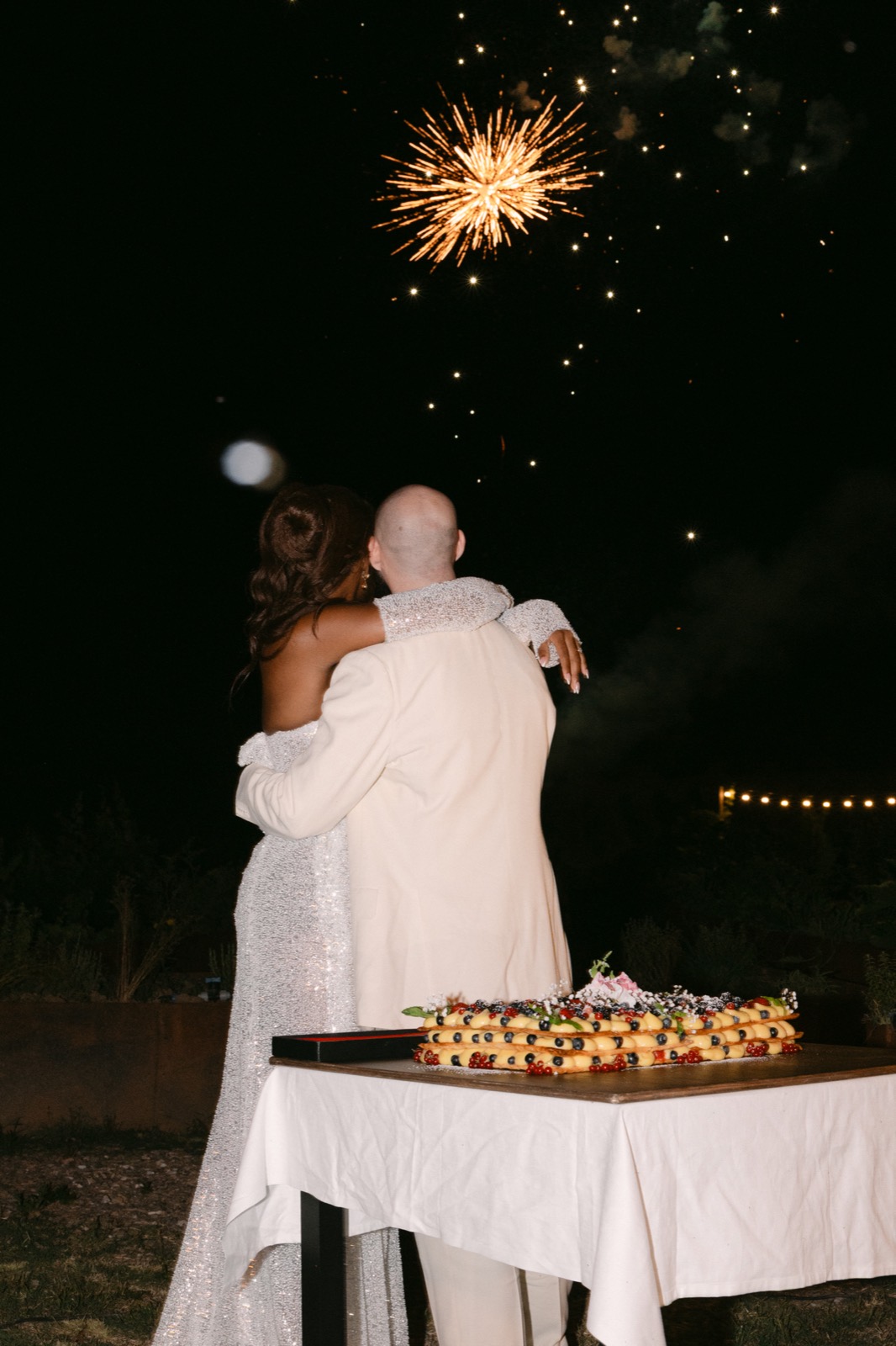 The couple embrace with their backs to the camera, watching fireworks beside a traditional Italian wedding cake